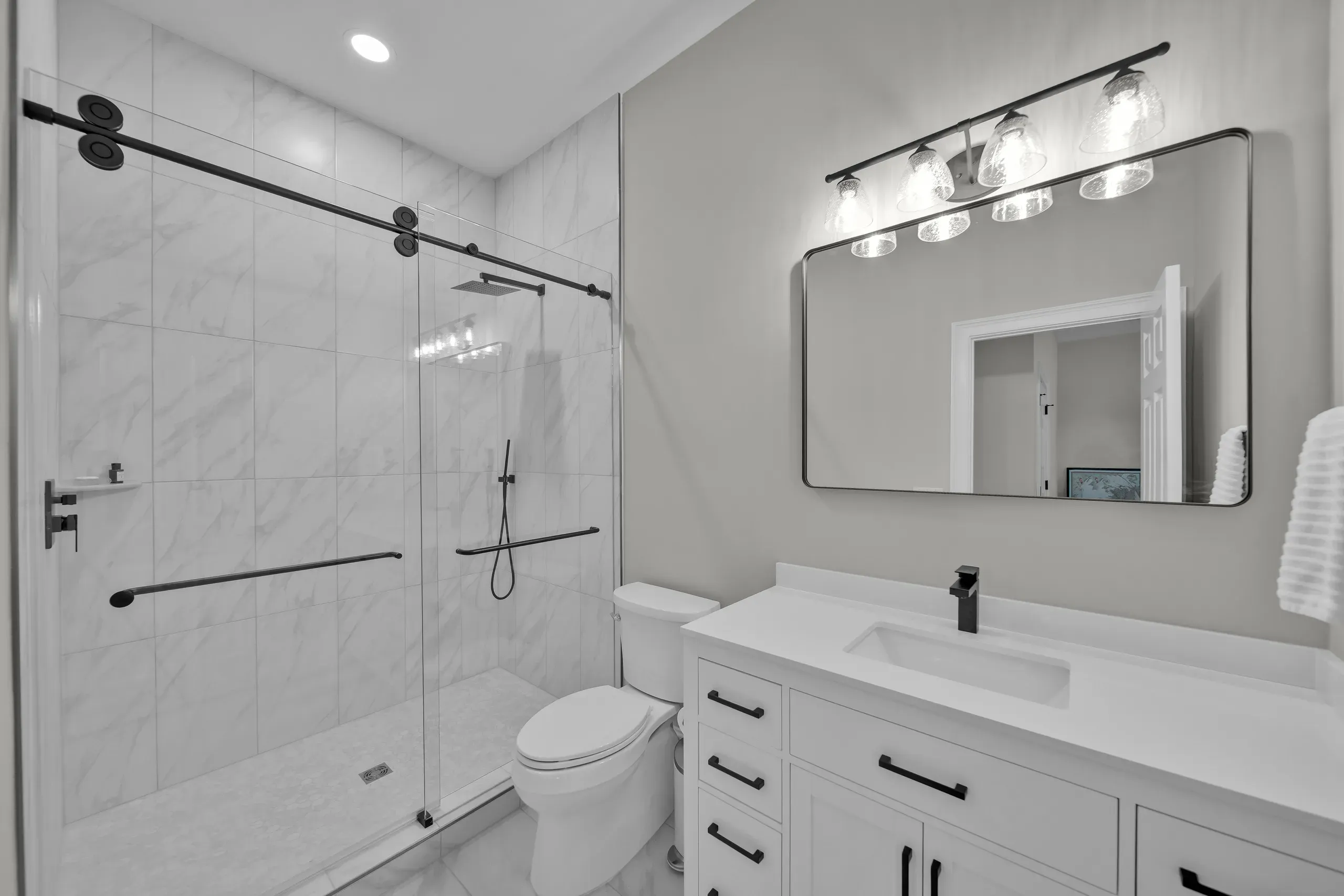 A remodeled bathroom features a marble-tiled walk-in shower with a black barn-door style glass enclosure and rainfall showerhead beside a white vanity with a rectangular sink and framed mirror.