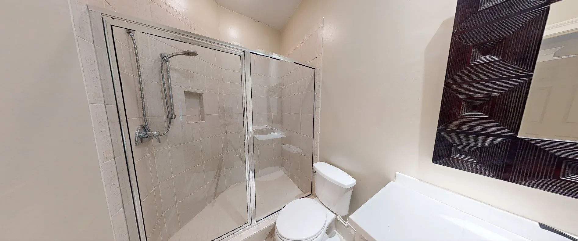 A bathroom features a framed glass shower enclosure with beige tile and a handheld showerhead, next to a white toilet and a dark wood-framed mirror above a vanity.