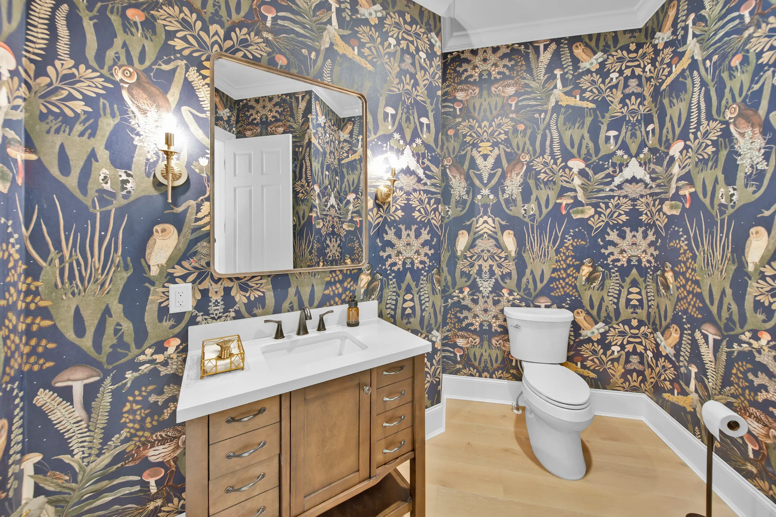 A powder room remodel features dramatic navy wallpaper printed with owls and mushrooms surrounding a wood vanity with white quartz countertop and a gold-framed mirror.