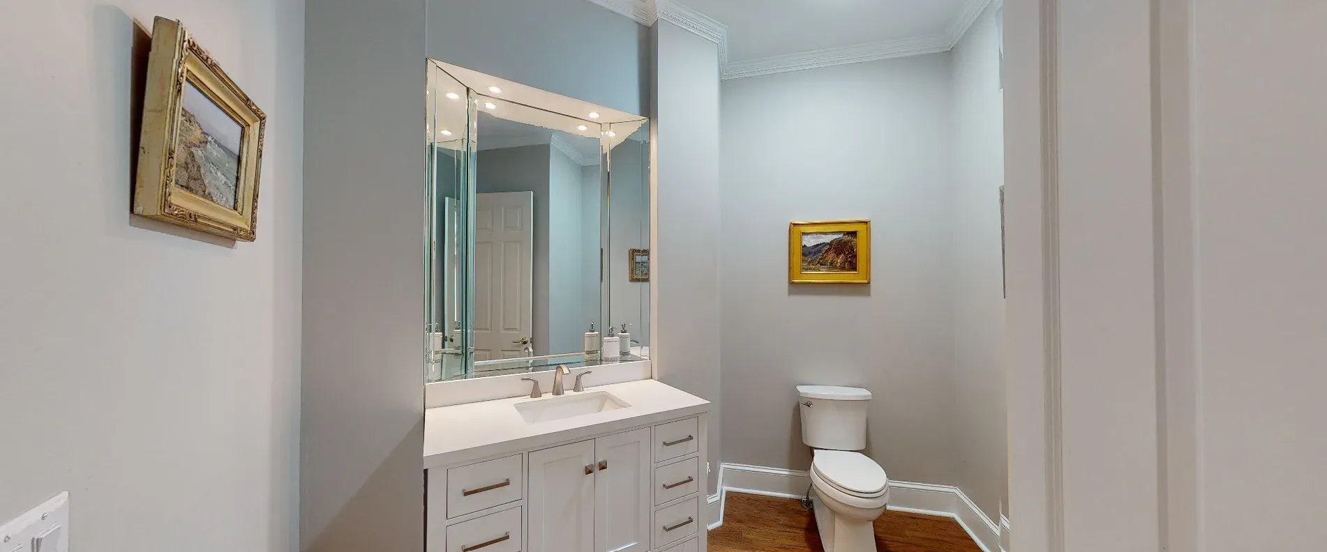 A remodeled powder room features a white shaker-style vanity with quartz countertop beneath a large angled mirror, framed artwork on soft gray walls, and a toilet set against a wall near the wood-look flooring.