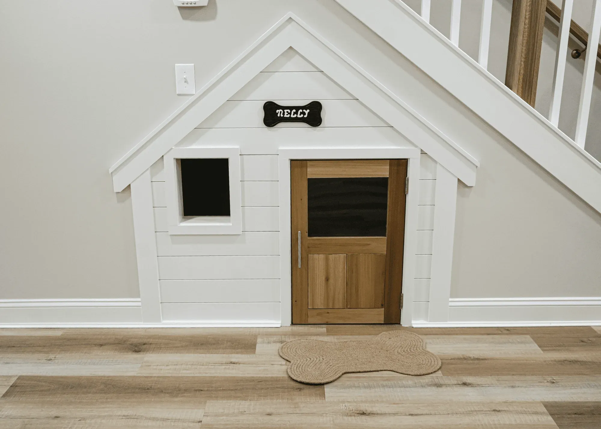 Built-in dog house under staircase with peaked roof, window, wood door, and bone-shaped rug