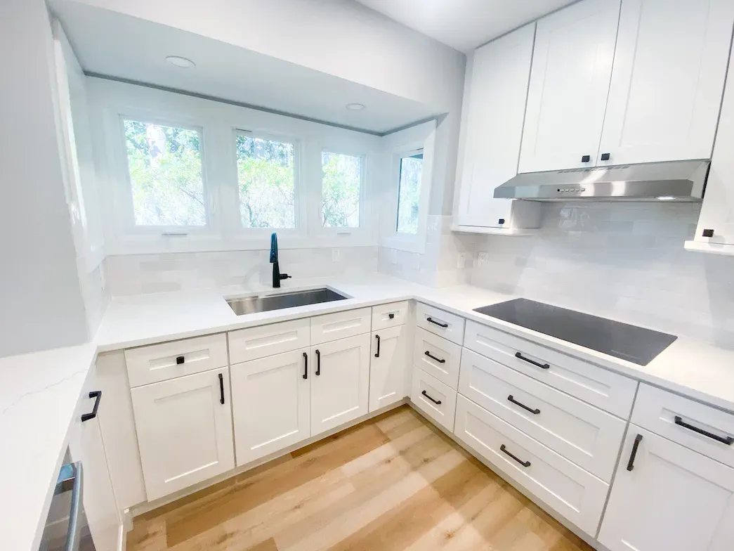 Kitchen with white cabinets, a corner sink below windows, and a cooktop under a range hood