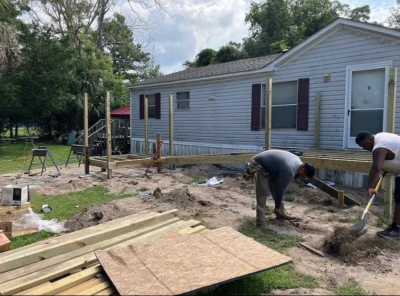 Workers building a wooden deck frame along a house, digging and shoveling dirt