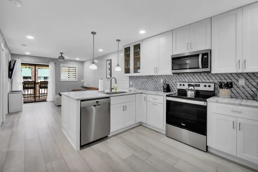 An open-concept kitchen remodel features white shaker cabinets, stainless steel appliances, marble countertops, and a diamond-patterned tile backsplash, opening onto a living area with sliding glass doors to a porch.