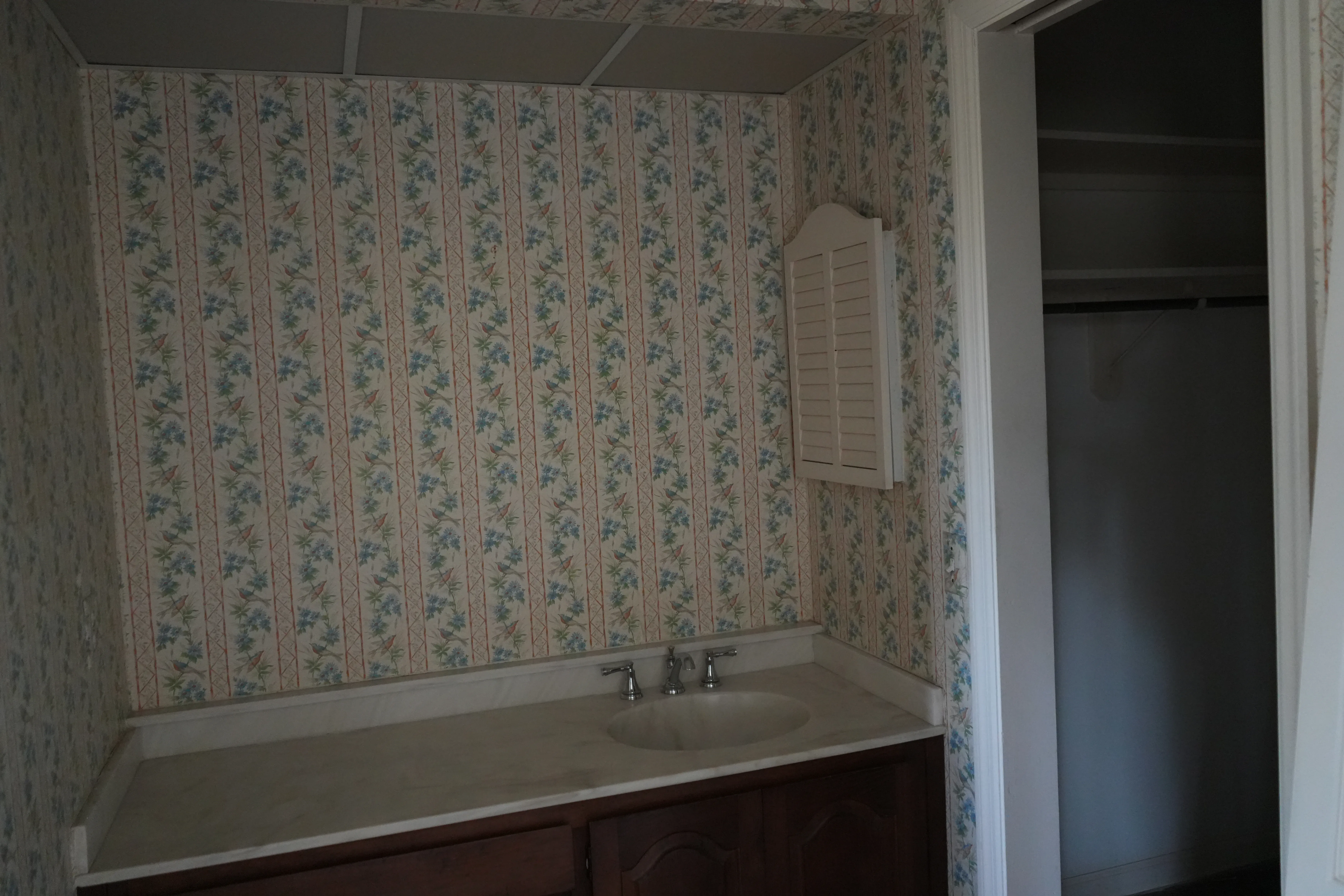 An outdated bathroom features floral wallpaper, a single sink vanity with dark wood cabinetry, and a small louvered shutter mounted on the wall beside an open closet.