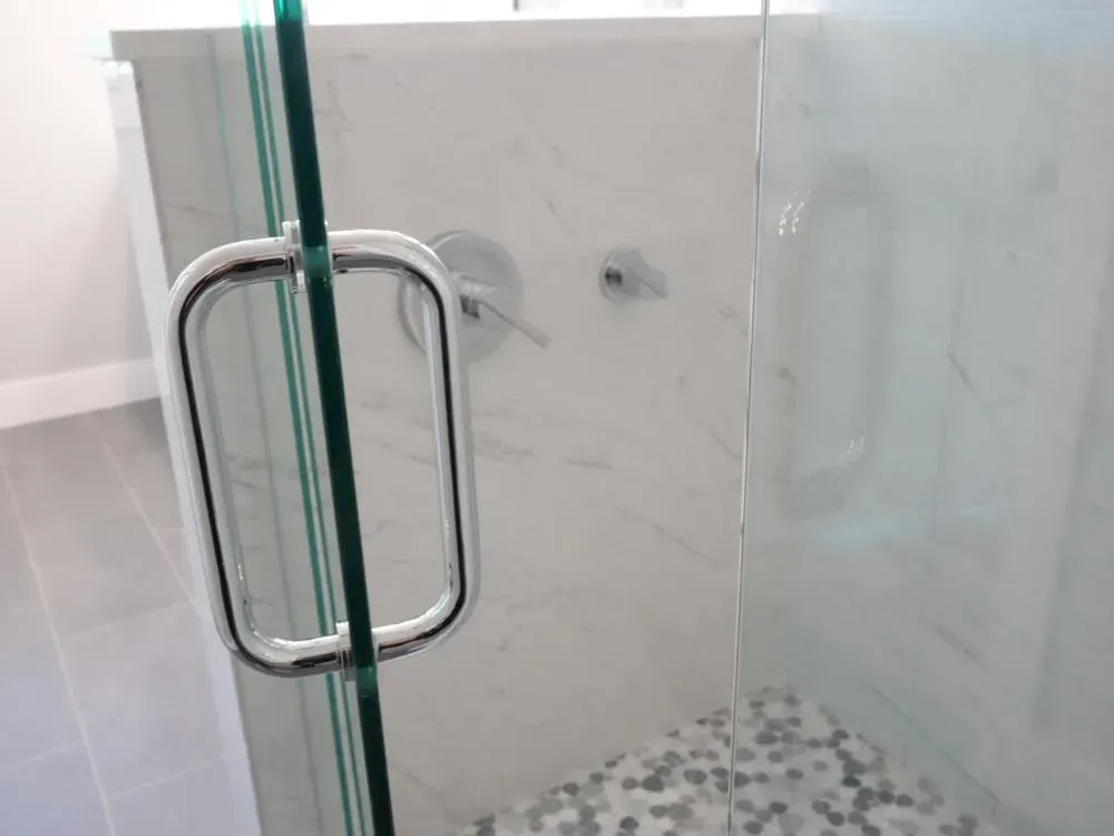 A frameless glass shower door with a chrome handle opens to a marble-tiled shower featuring a pebble mosaic floor and chrome fixtures.