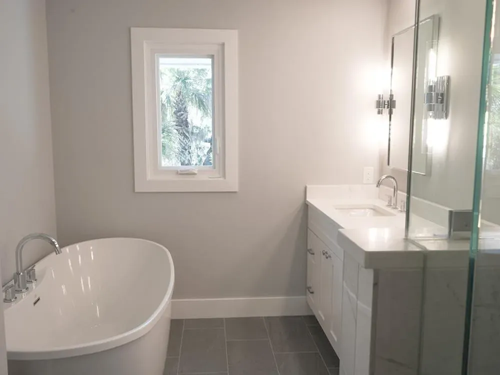 A remodeled bathroom features a freestanding oval soaking tub beside a window with a palm tree view, and a white vanity with a marble countertop and framed mirror lit by wall sconces.