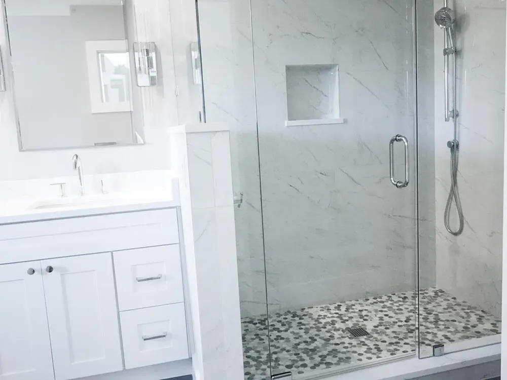 A remodeled bathroom features a white vanity with quartz countertop beside a frameless glass shower enclosure with marble-look tile walls and a pebble mosaic floor.