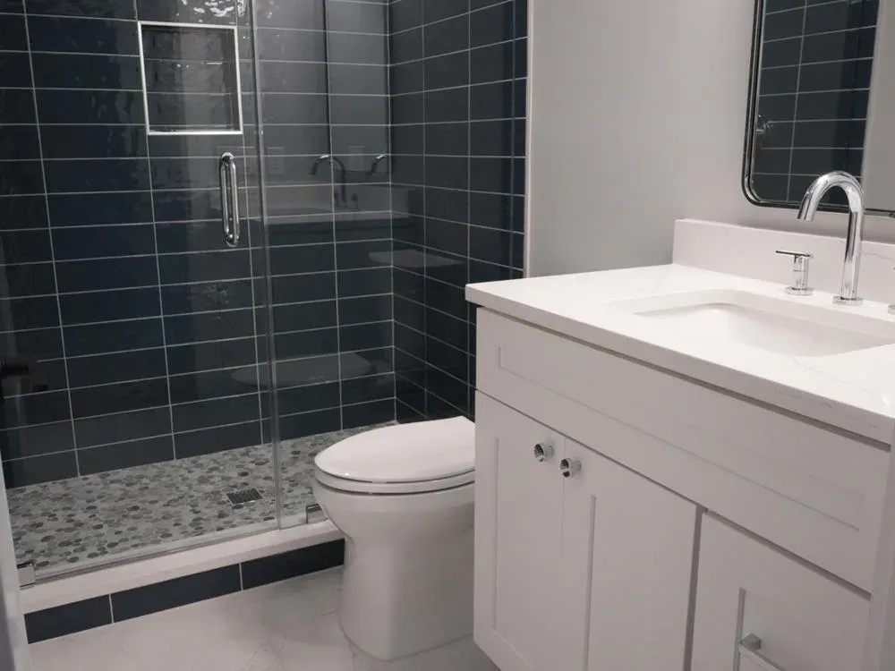 A remodeled bathroom features a glass-enclosed shower with dark navy subway tile and pebble flooring beside a white vanity with a quartz countertop and chrome fixtures.