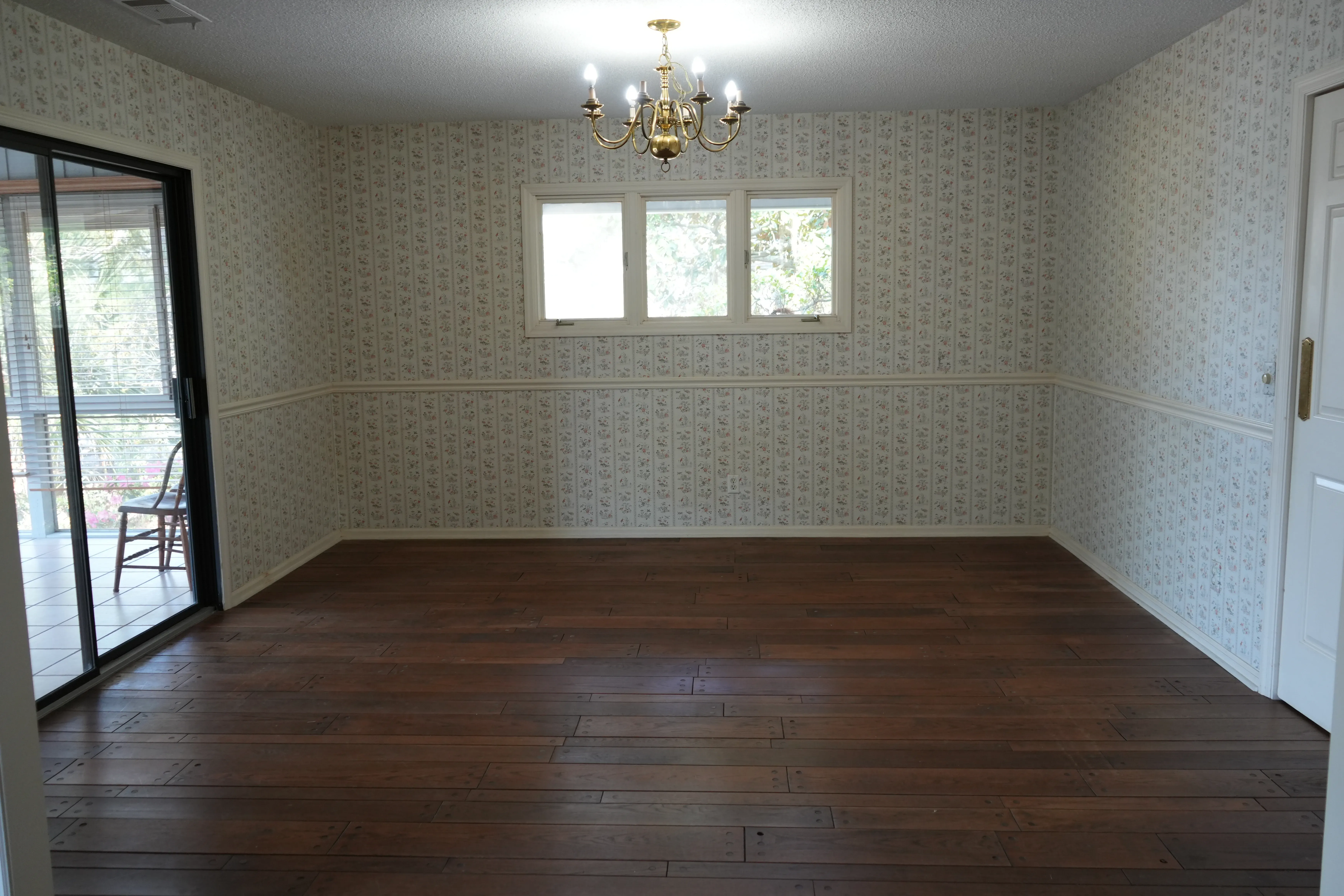 An empty dining room with dark hardwood flooring, floral wallpaper, a brass chandelier, and a sliding glass door leading to a screened porch.