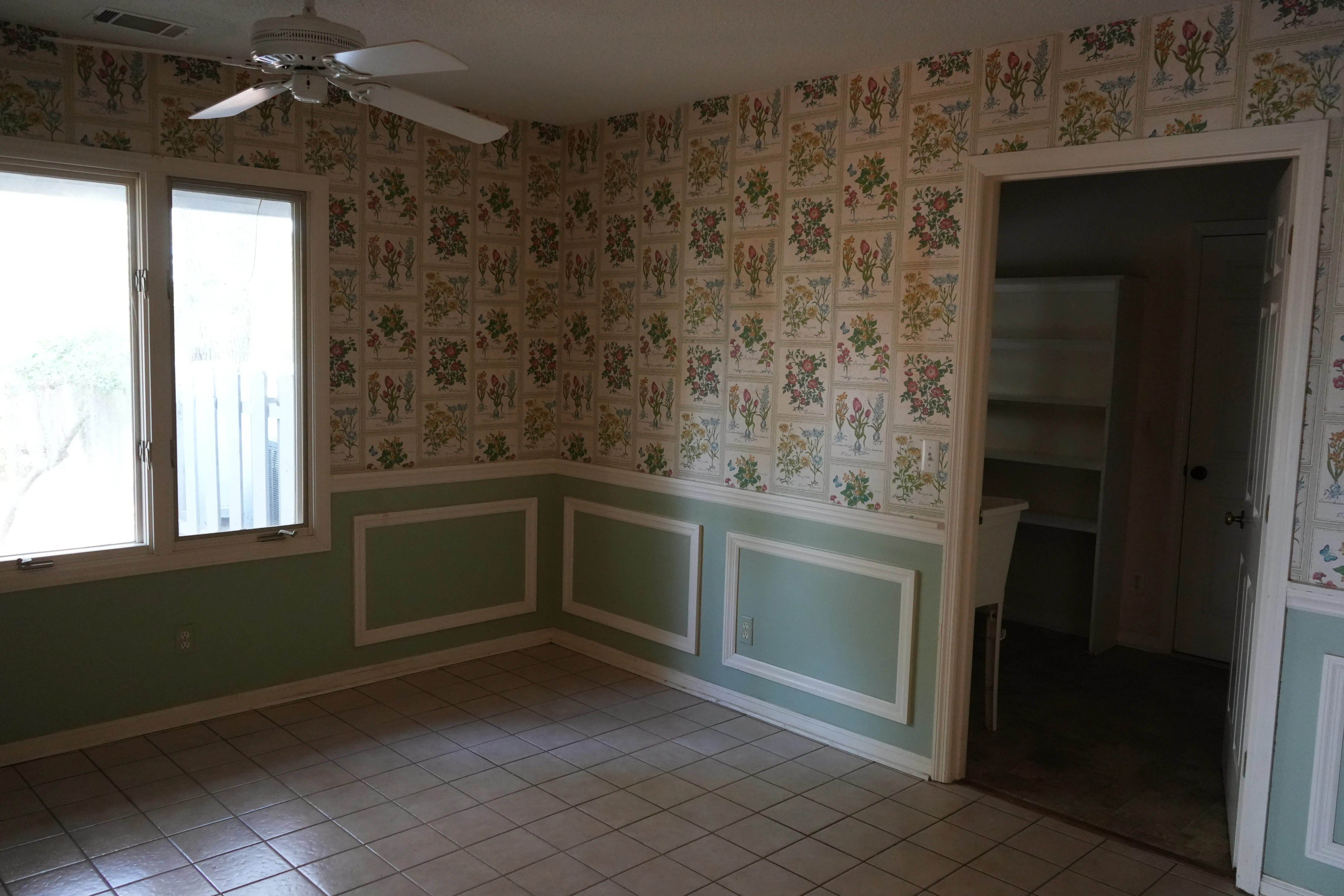 An empty dining room before renovation shows floral wallpaper above green wainscoting, a tile floor, a ceiling fan, and a doorway leading to a small built-in shelving nook.