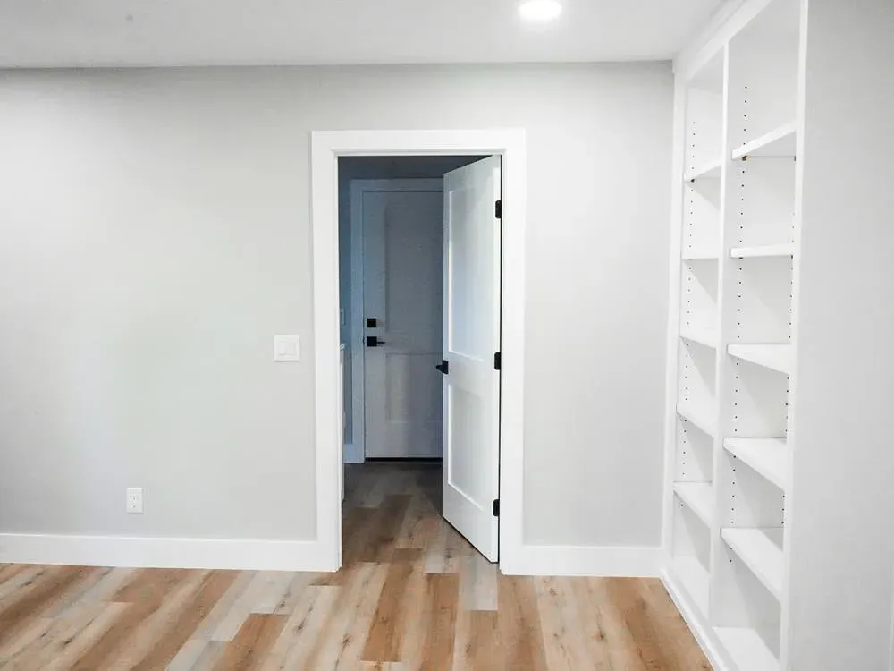 An empty newly remodeled room with light wood flooring, white built-in shelving, and an open door leading to a hallway.