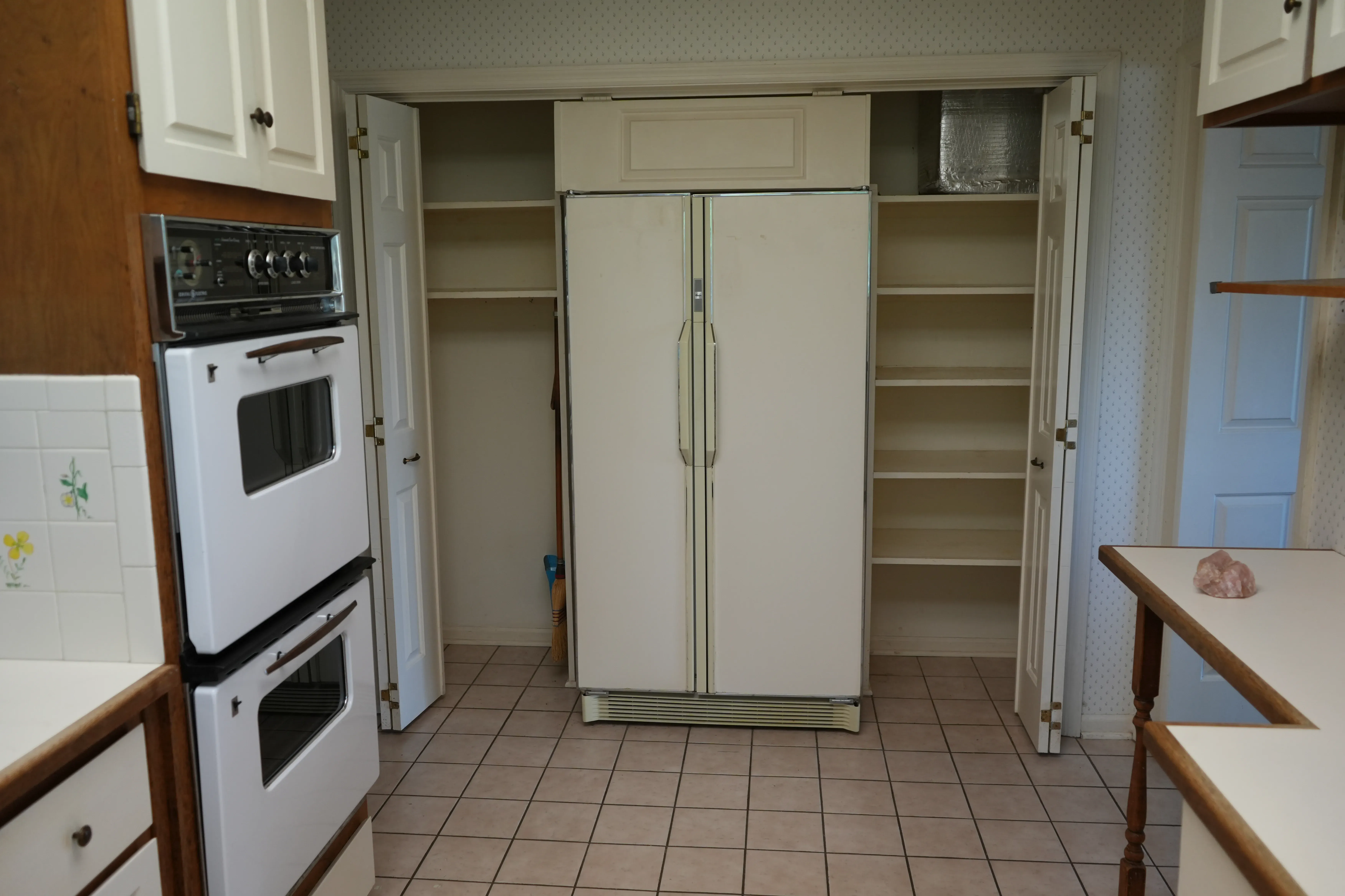 An outdated kitchen shows a white double wall oven, a bisque-colored side-by-side refrigerator flanked by open pantry closets, and worn tile flooring before renovation.