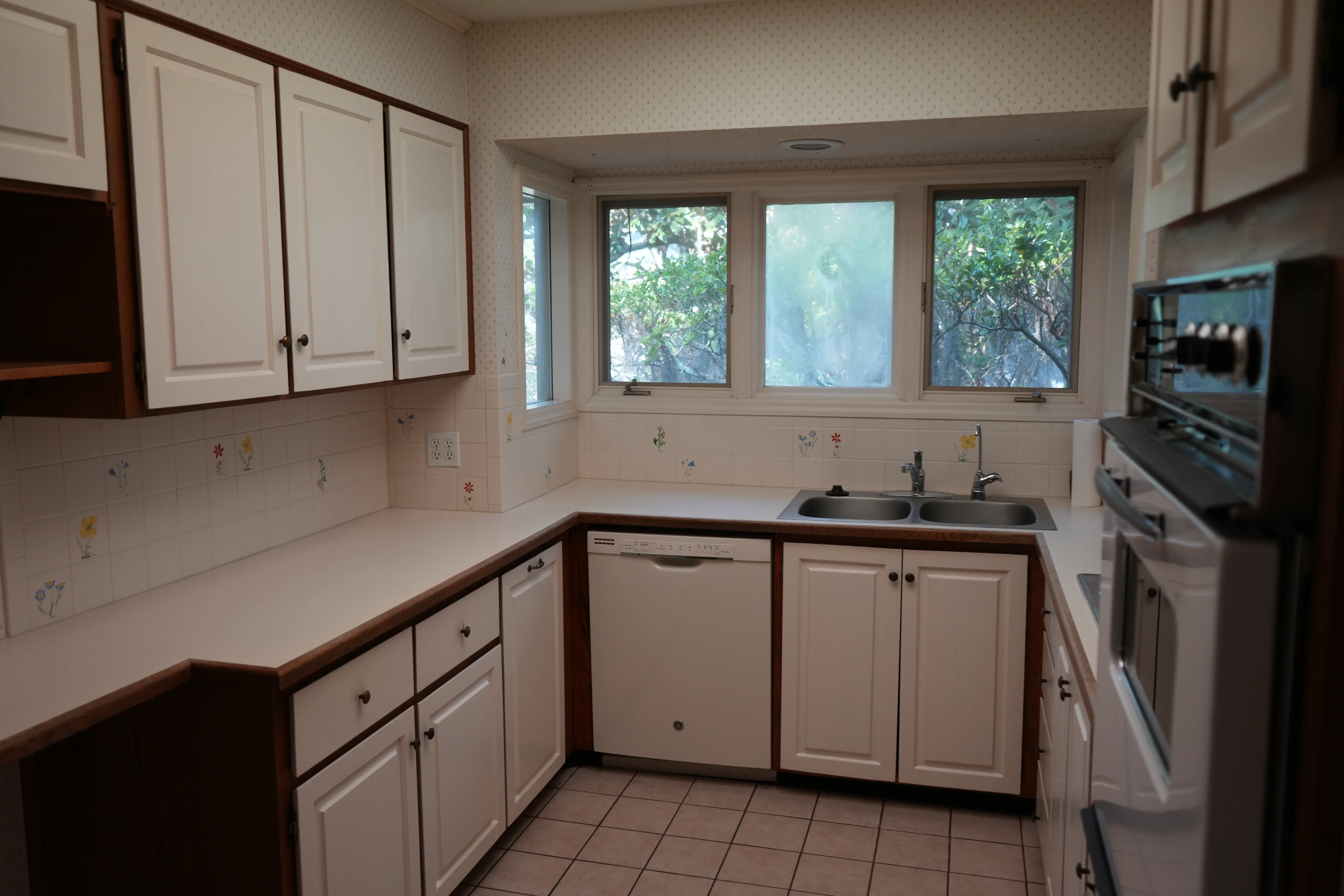 An outdated kitchen features white cabinets with wood trim, tile flooring, a stainless double sink beneath a row of windows, a white dishwasher, and a wall oven with an adjacent stainless microwave.