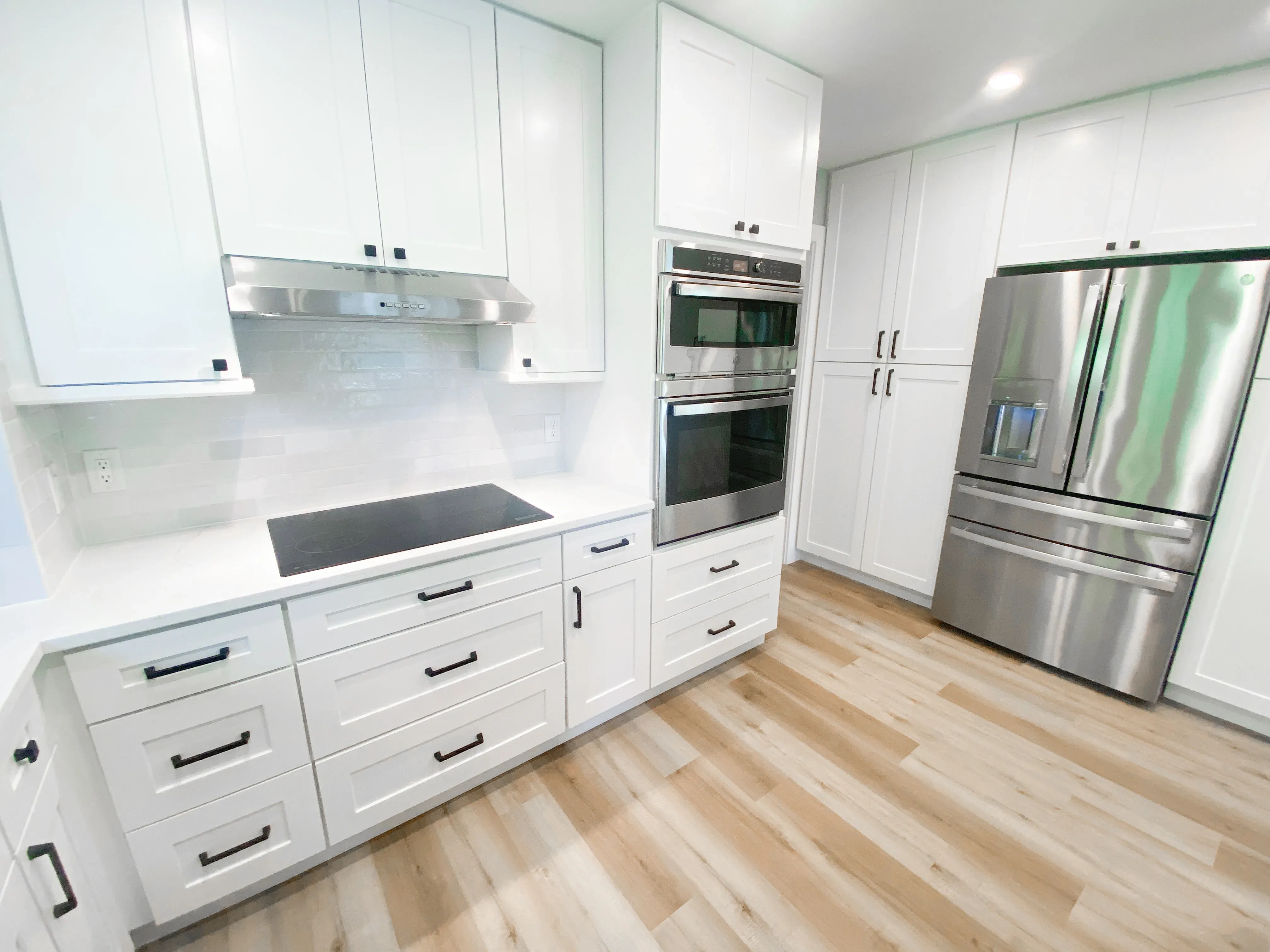 A remodeled kitchen features white shaker cabinets, a stainless steel french-door refrigerator, a built-in double oven with microwave, and a black induction cooktop set into white countertops above light wood flooring.