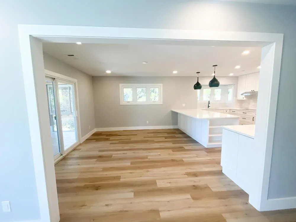 An open, newly remodeled space shows a white kitchen with a large island and dark pendant lights flowing into an adjoining room with light wood flooring and sliding glass doors.