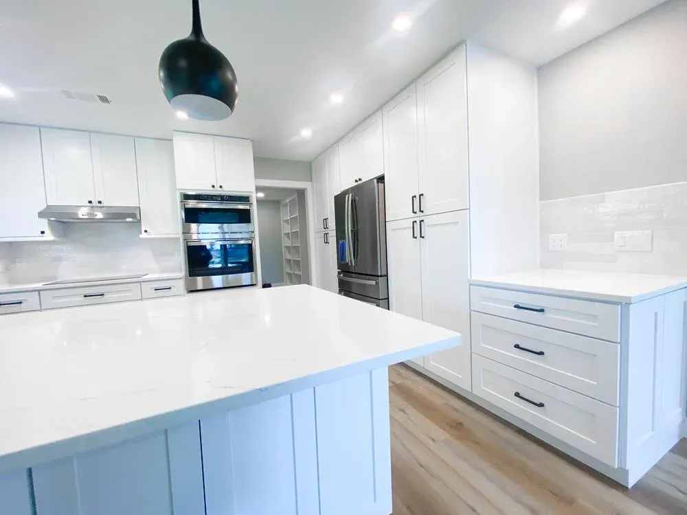 A bright white kitchen features a large quartz-topped island, stainless steel double wall ovens and refrigerator, floor-to-ceiling storage cabinets, and light wood flooring under recessed and pendant lighting.