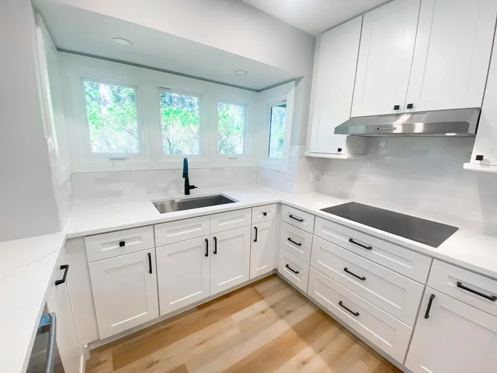 A remodeled kitchen features white shaker cabinets with black hardware, a quartz countertop with an undermount sink, a black induction cooktop with stainless hood, and a bay window overlooking greenery.
