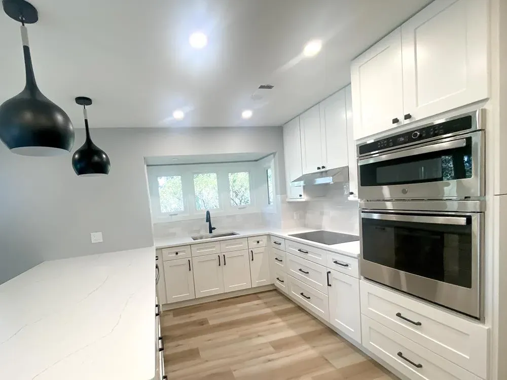 A remodeled kitchen features white shaker cabinets, quartz countertops, stainless steel double wall ovens, a built-in microwave, and a bay window over the sink that overlooks trees, with dark pendant lights hanging above a wood-floored island area.