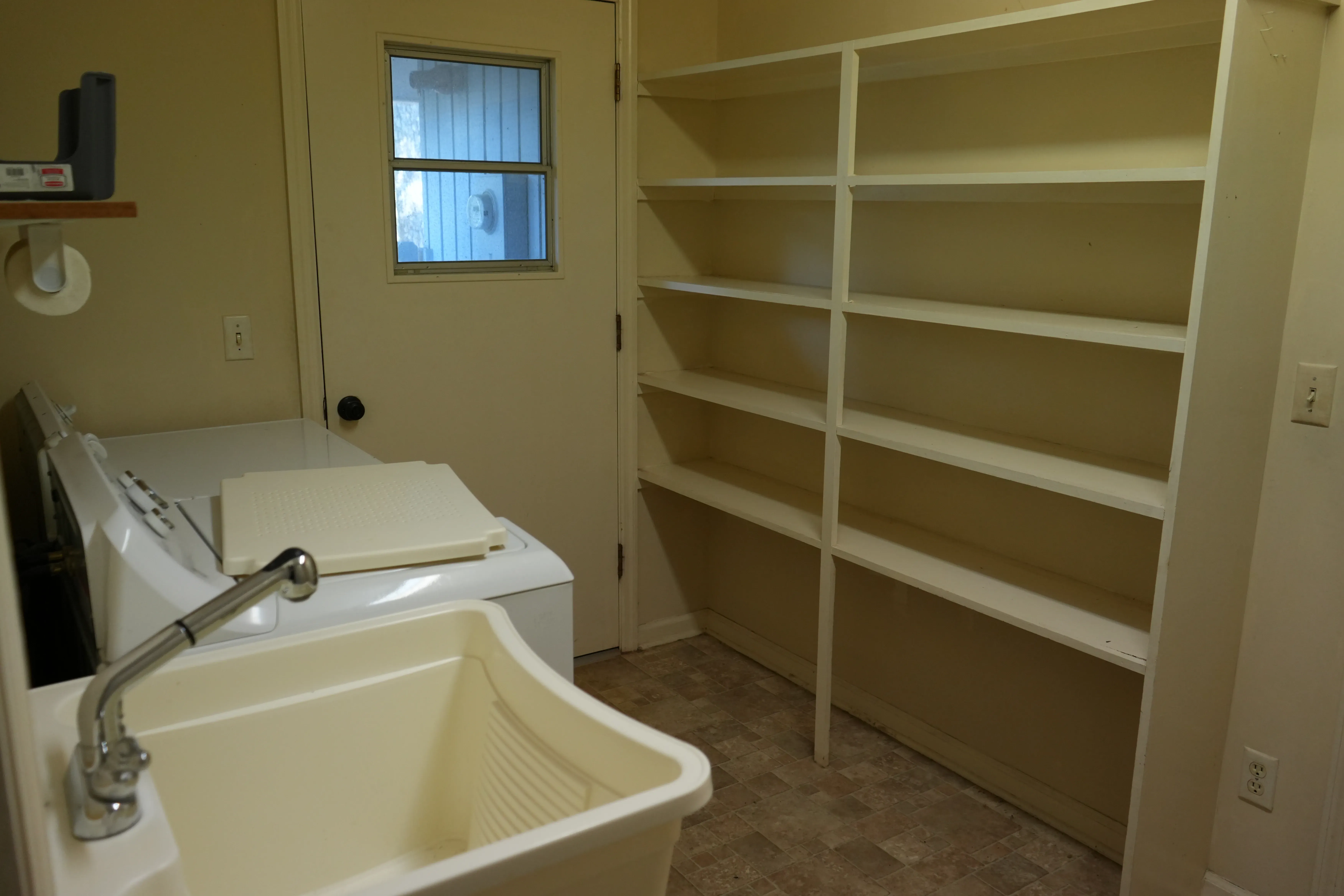 An outdated laundry room with a utility sink, washing machine, an exterior door with a small window, and tall built-in white shelving along the back wall.