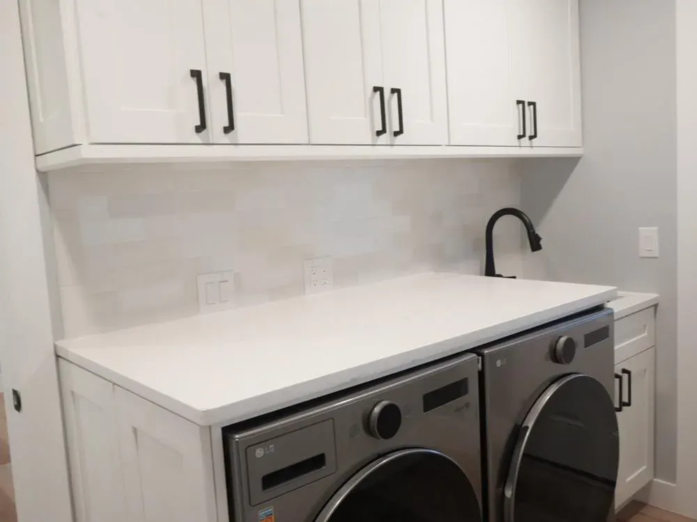 A remodeled laundry room features white shaker cabinets with black hardware, a quartz countertop with an integrated sink and matte black faucet, and a stacked front-loading washer and dryer set beneath the counter.