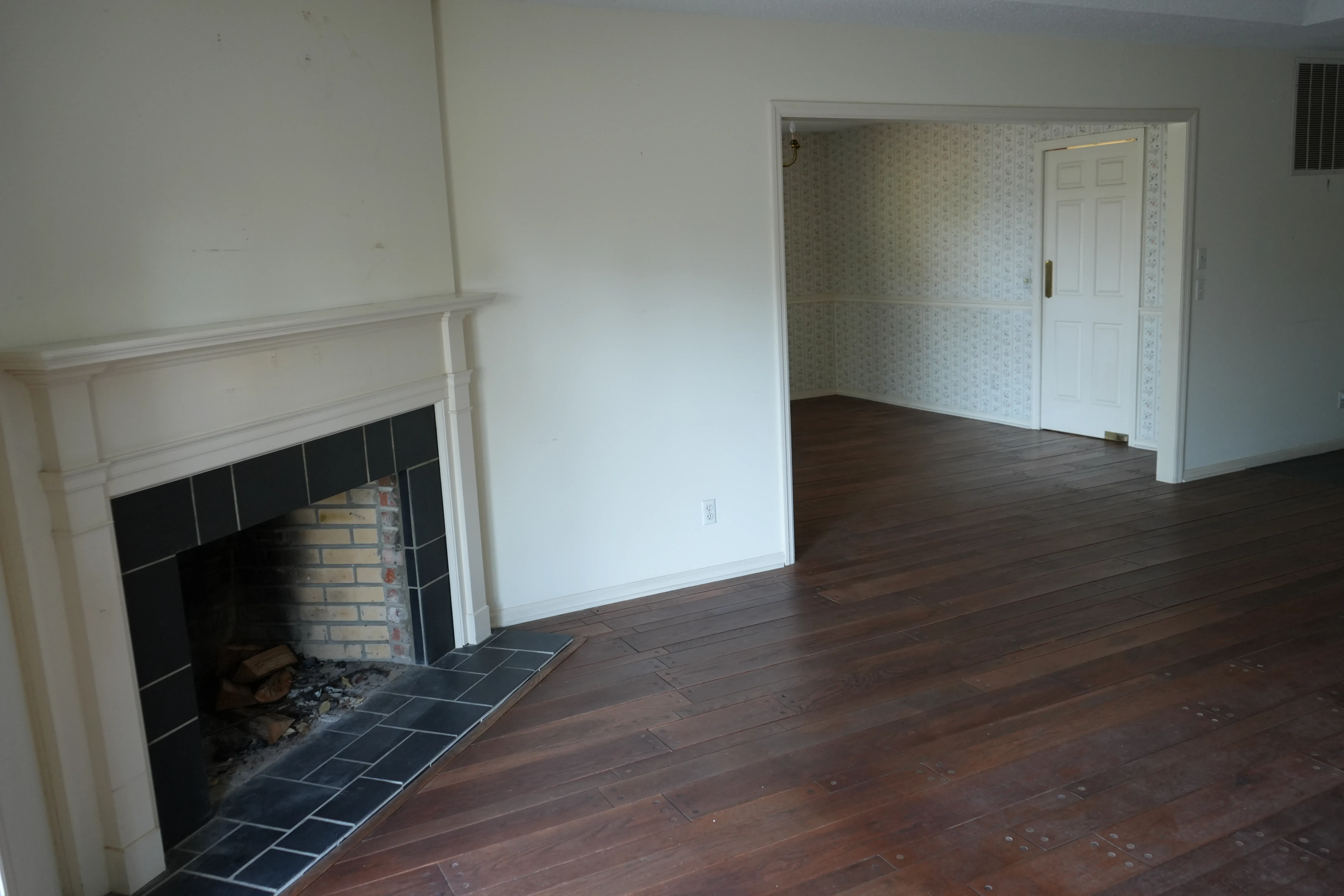 An empty living room before renovation shows a dark hardwood floor, a white mantel with a black tile surround around the fireplace, and an open doorway leading to an adjoining room with floral wallpaper.