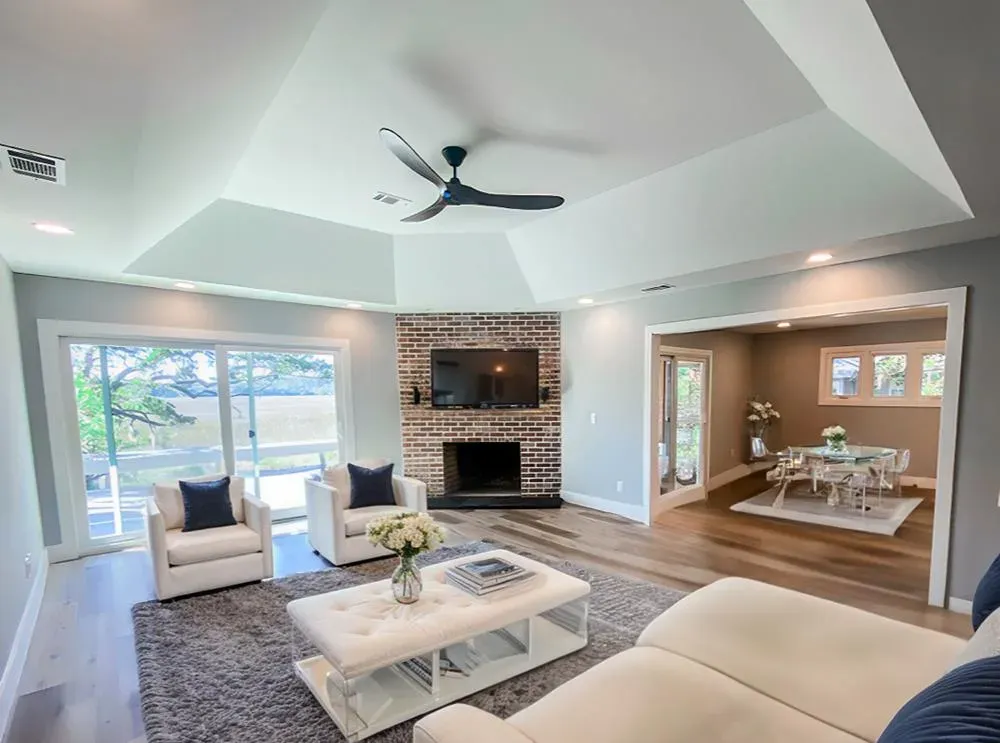 A living room with a tray ceiling and ceiling fan features a brick fireplace with a mounted television, cream armchairs, a tufted ottoman, and a sliding glass door leading to a water view, with a dining area visible beyond.