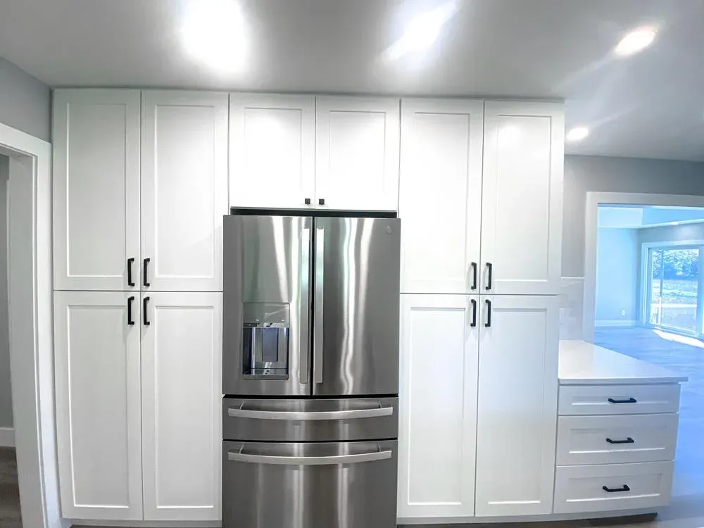 A stainless steel French-door refrigerator with bottom freezer drawers is flanked by tall white shaker-style pantry cabinets with black hardware, with a bright open living area visible through a doorway to the right.