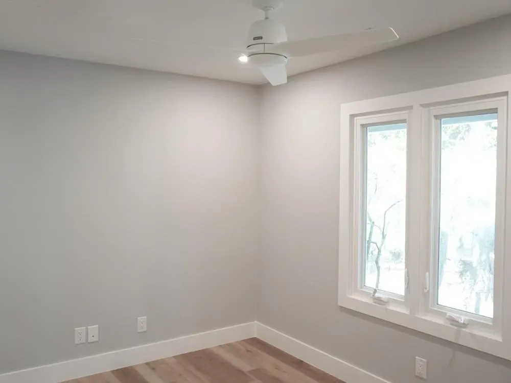 A newly finished empty room with light gray walls, white trim, a white ceiling fan, and two large windows above a wood-look floor.
