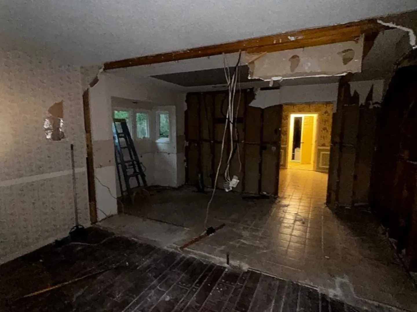 An interior space mid-demolition with exposed wall studs, hanging wires, a step ladder, and a tiled hallway leading to a distant room lit in yellow light.