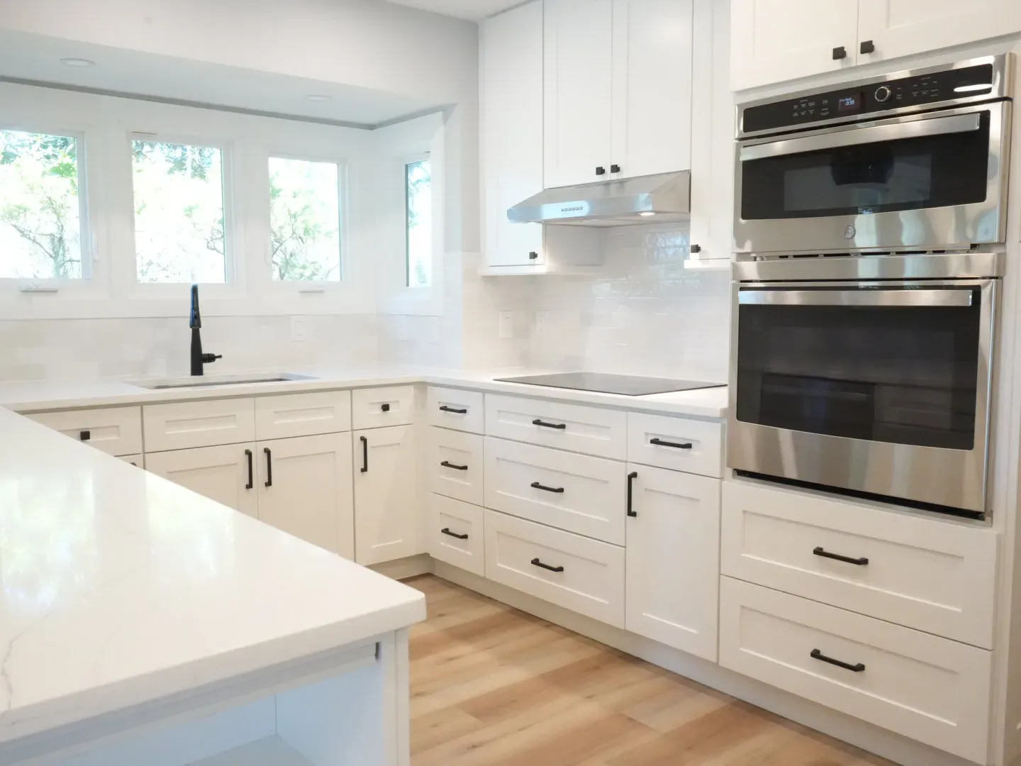A remodeled kitchen features white shaker cabinets with black hardware, a quartz countertop, stainless double wall ovens, and a bay window above the sink overlooking trees.