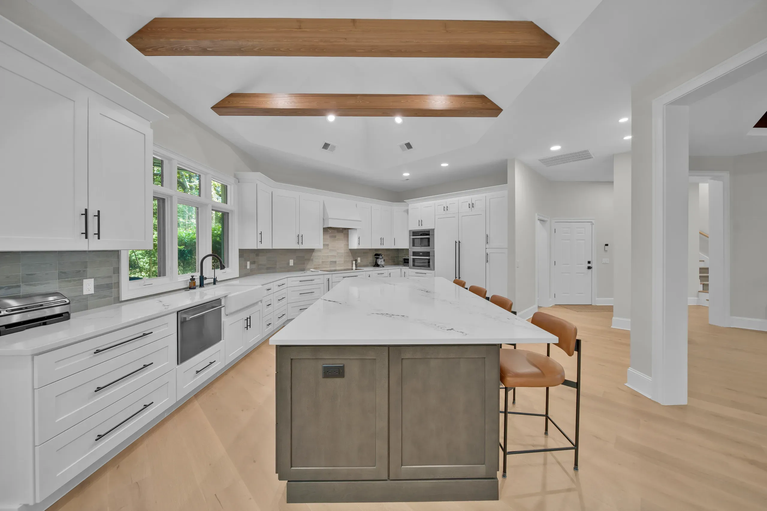 A bright kitchen remodel features white shaker cabinets, a large quartz-topped island with a gray wood base and leather counter stools, exposed wood ceiling beams, and light hardwood floors leading into an open hallway.