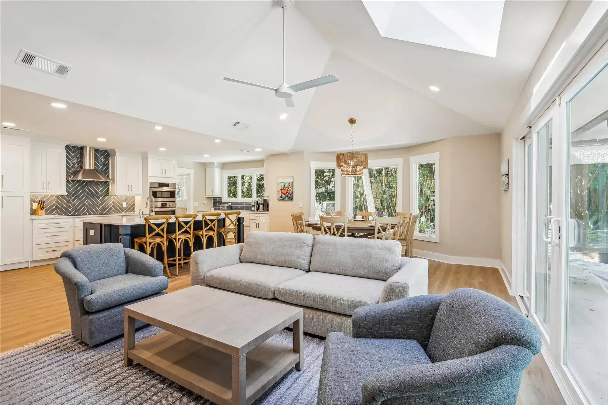 An open-concept living area with gray sofas and armchairs around a wooden coffee table flows into a white kitchen with a dark herringbone tile backsplash and island seating, all beneath a vaulted ceiling with a skylight and ceiling fan.