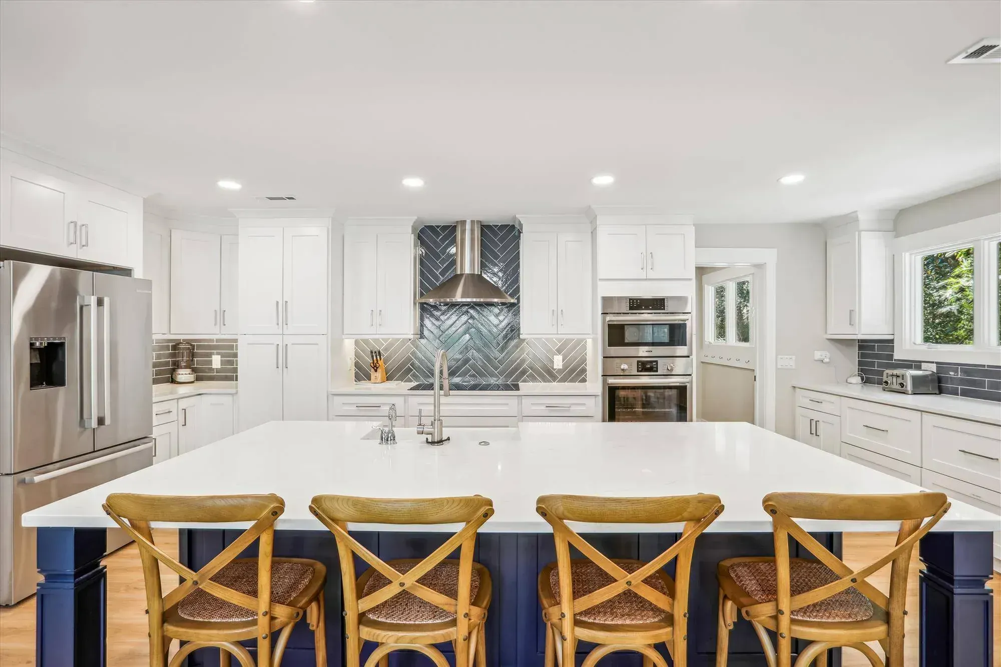 A bright white kitchen features a large navy blue island with wooden bar stools, quartz countertops, and a herringbone tile backsplash behind the stove and stainless steel range hood.