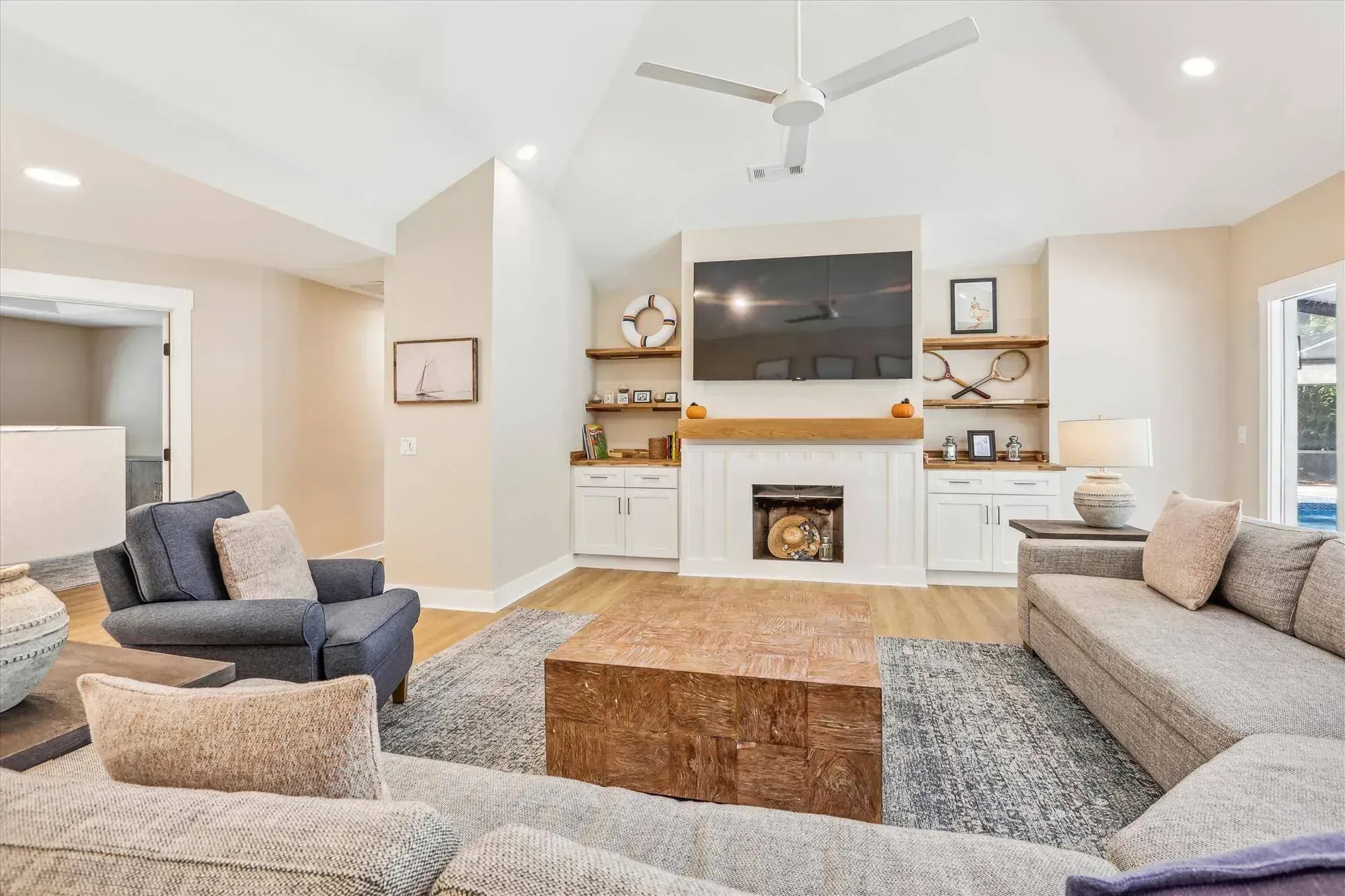A vaulted living room features a white fireplace flanked by built-in wood-topped shelving and cabinetry, with a mounted television above the mantel and light wood flooring topped by a gray patterned rug and sectional seating.
