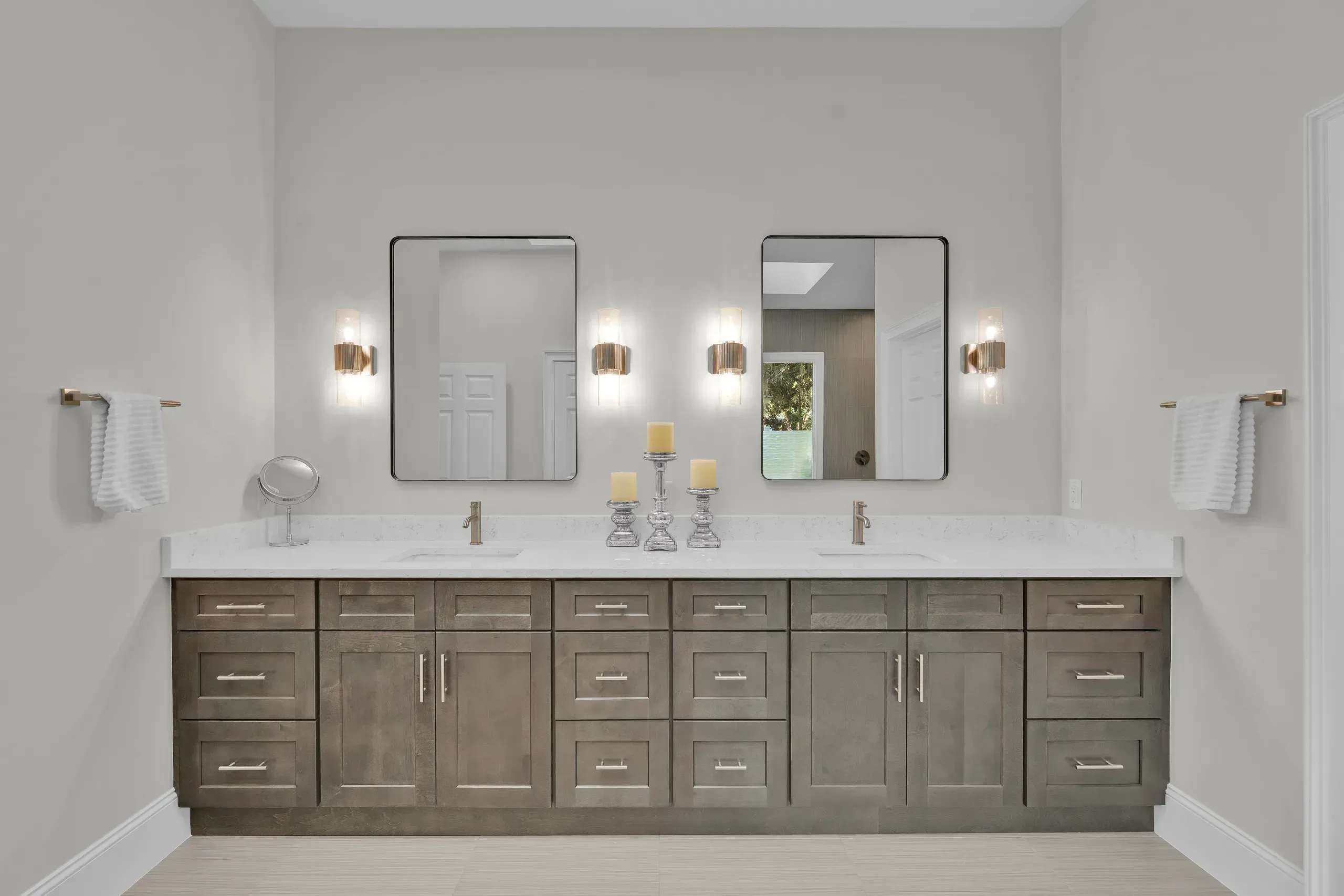 A long double-sink bathroom vanity with dark wood cabinetry, white countertops, two rectangular mirrors, and gold wall sconces flanking each mirror.