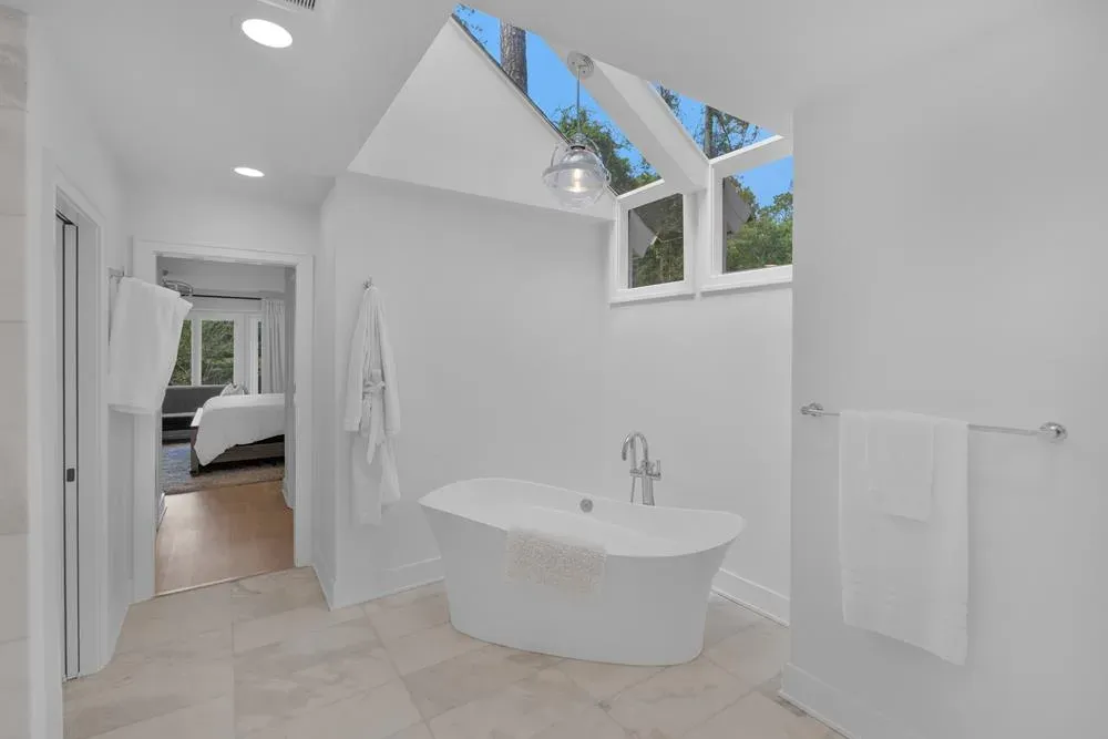A freestanding white soaking tub sits beneath a peaked skylight in a bright bathroom that opens directly into the adjoining bedroom.