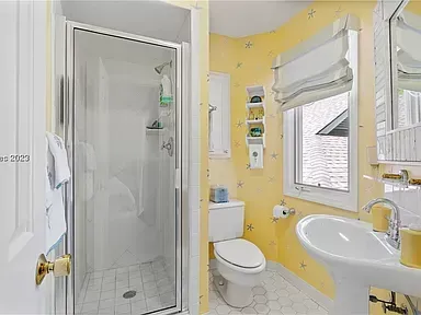 A bright yellow bathroom features a glass-enclosed shower, a pedestal sink, a toilet, and a window with a fabric shade above small corner shelves.