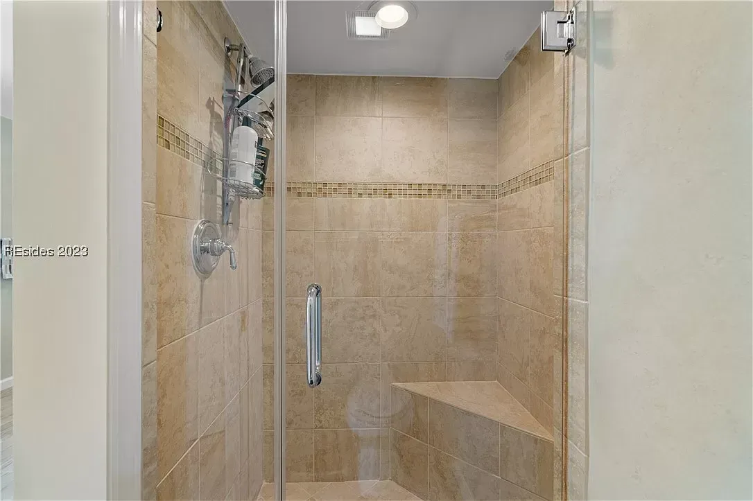 A glass-enclosed shower features beige tile walls with a decorative mosaic accent band, a built-in corner seat, and a chrome handheld showerhead with grab bar.