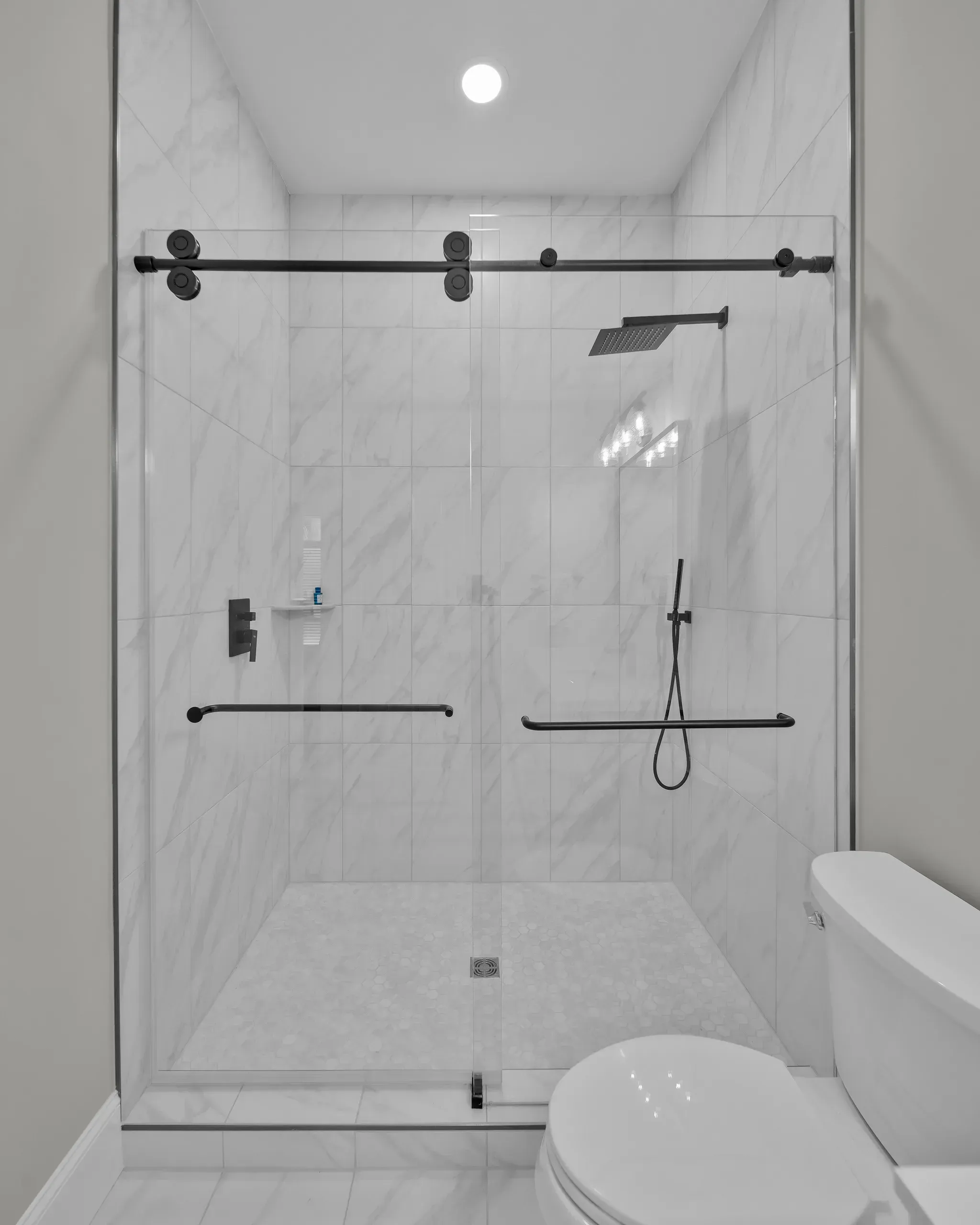A frameless glass shower enclosure with black hardware and marble-look tile walls features a rain showerhead, handheld sprayer, and black grab bars above a hexagon tile floor.