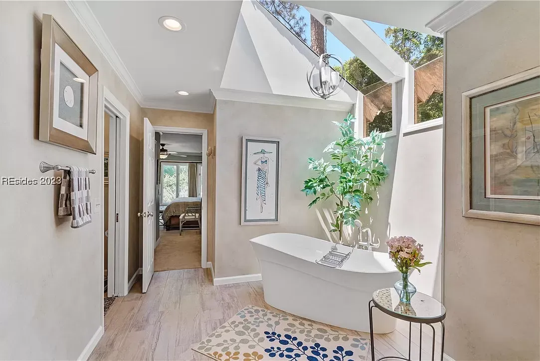 A freestanding soaking tub sits beneath a vaulted skylight in a bright primary bathroom with light wood-look flooring and a view through to the bedroom beyond.