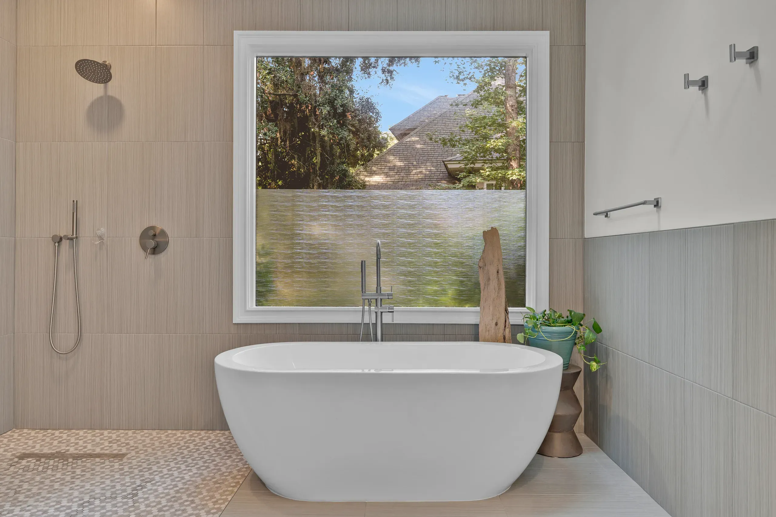 A freestanding white oval soaking tub sits beneath a large window with a frosted lower pane, positioned between a tiled walk-in shower and a decorative wood accent piece with a potted plant.