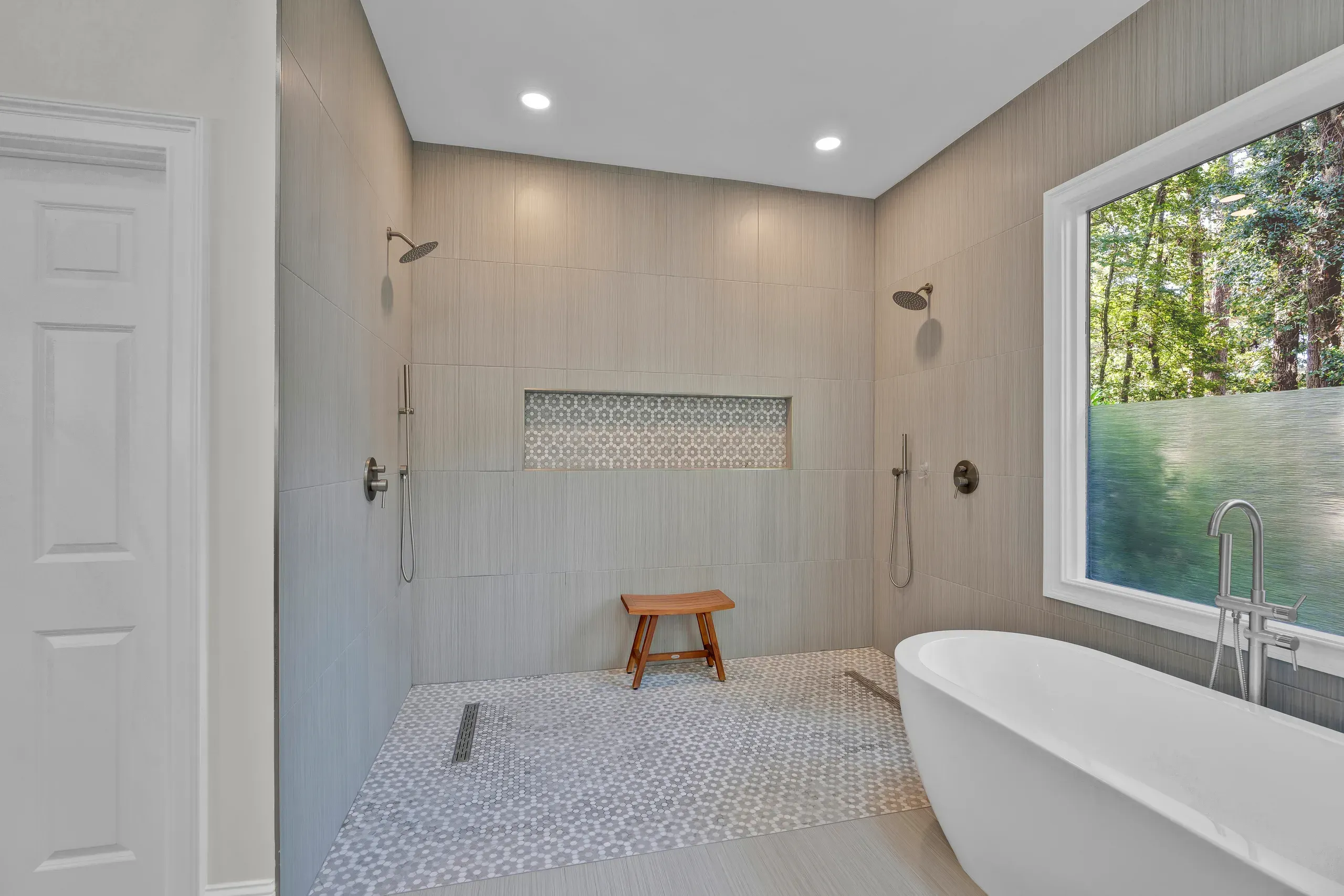 A spacious walk-in shower with dual rain showerheads and hexagon tile flooring sits beside a freestanding soaking tub near a window overlooking trees.