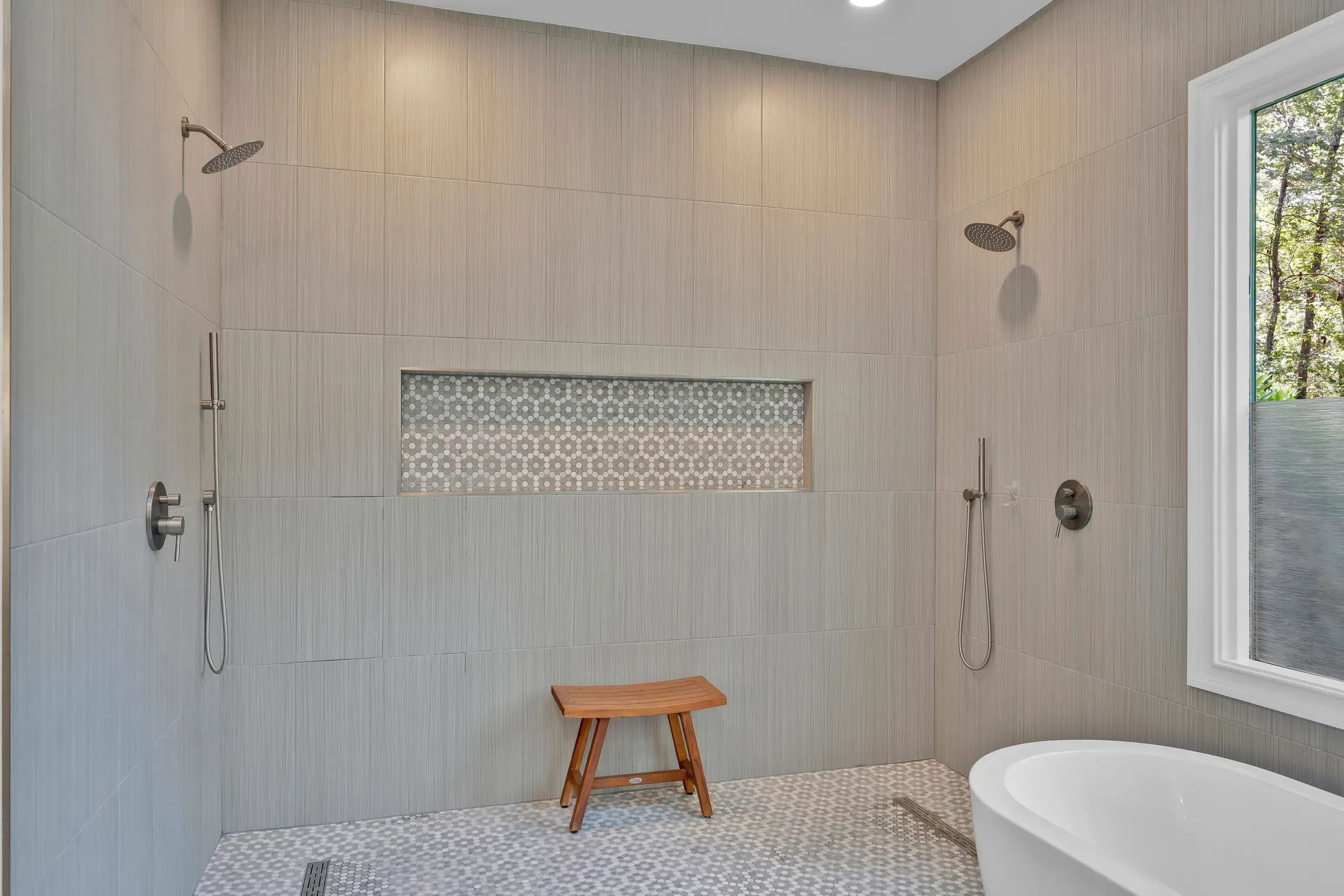 A spacious walk-in shower features two rain showerheads with handheld sprayers on opposite tiled walls, a penny-tile floor, a wooden teak bench, a mosaic tile niche, and a freestanding tub beside a window in the corner.