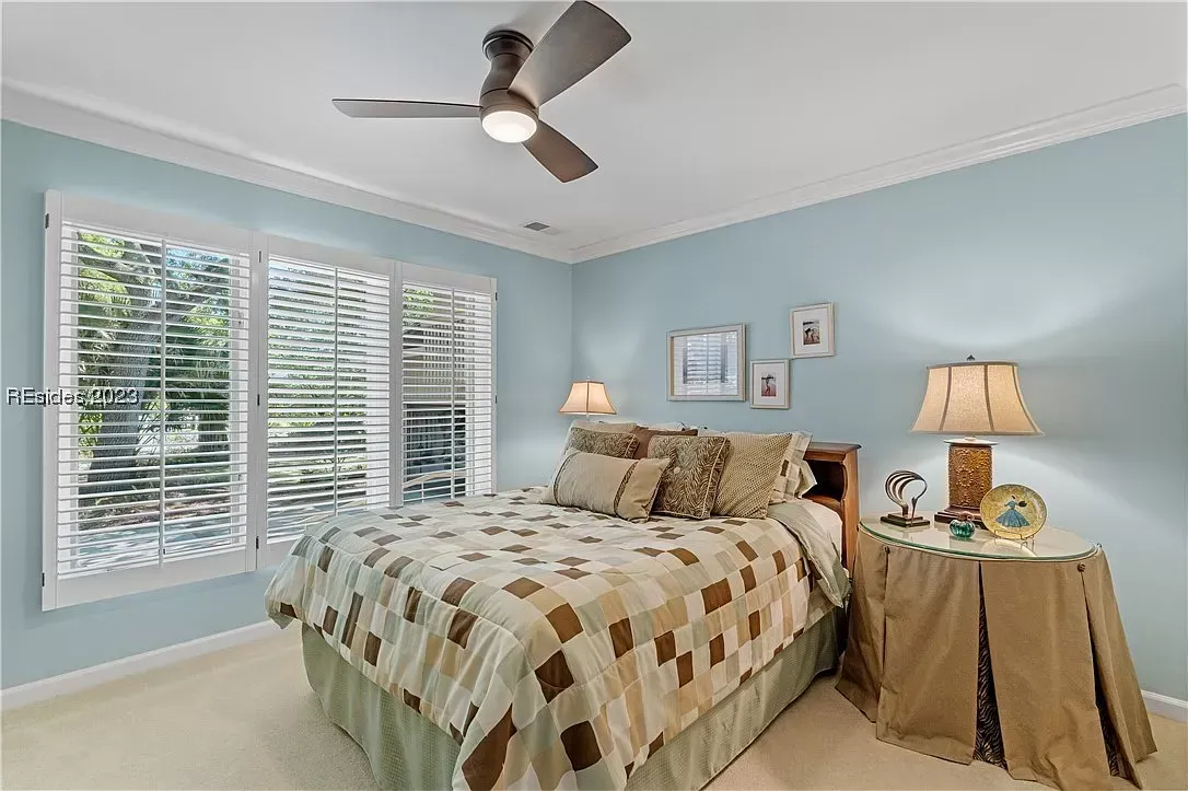 A light blue bedroom features a checkered quilted bedspread, plantation shutters on the windows, crown molding, and a ceiling fan overhead.