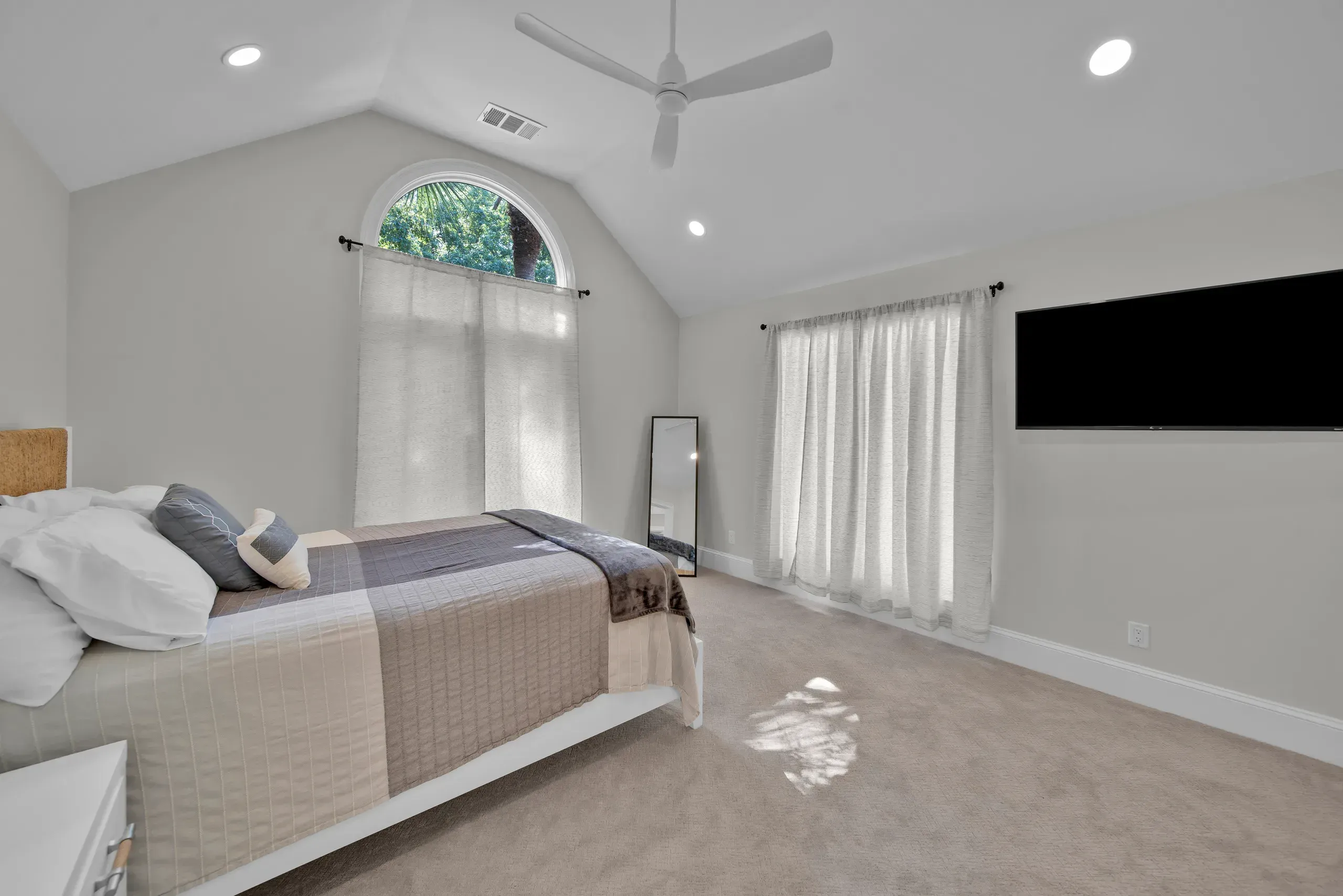 A bright bedroom with a vaulted ceiling, ceiling fan, and an arched window framed by sheer curtains above a bed with taupe and gray linens.