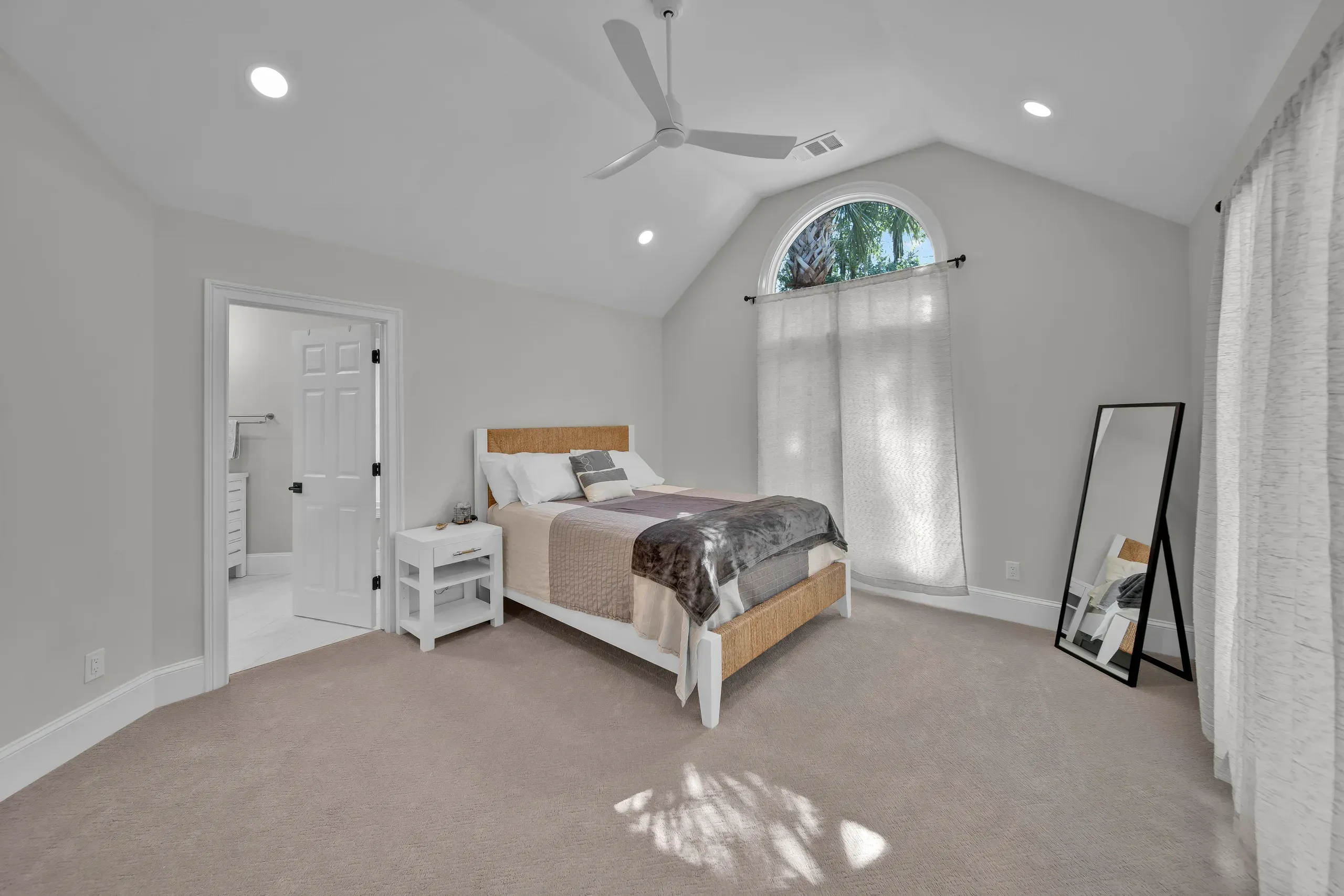 A spacious bedroom with a vaulted ceiling and ceiling fan features a woven headboard bed, a tall arched window with sheer curtains, a full-length mirror, and carpeted flooring leading to an adjoining bathroom.