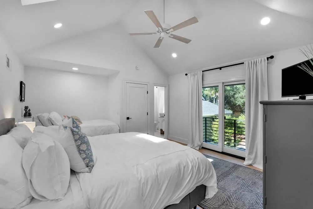 A bright bedroom addition with a vaulted ceiling and ceiling fan features two white beds, a sliding glass door leading to a balcony, and a view of trees through sheer curtains.