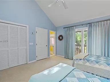 A light-filled bedroom with pale blue walls and a vaulted ceiling features white louvered closet doors, a glass door leading to an outdoor balcony, and beds dressed in blue and white quilts.