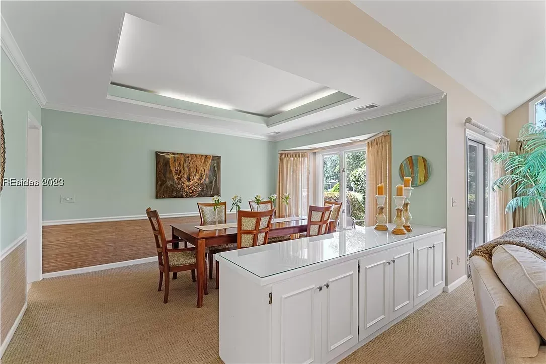 A dining room with sage green walls and a tray ceiling features a wooden table with six chairs beside a white built-in buffet cabinet with a marble top, opening onto a bay window with a garden view.